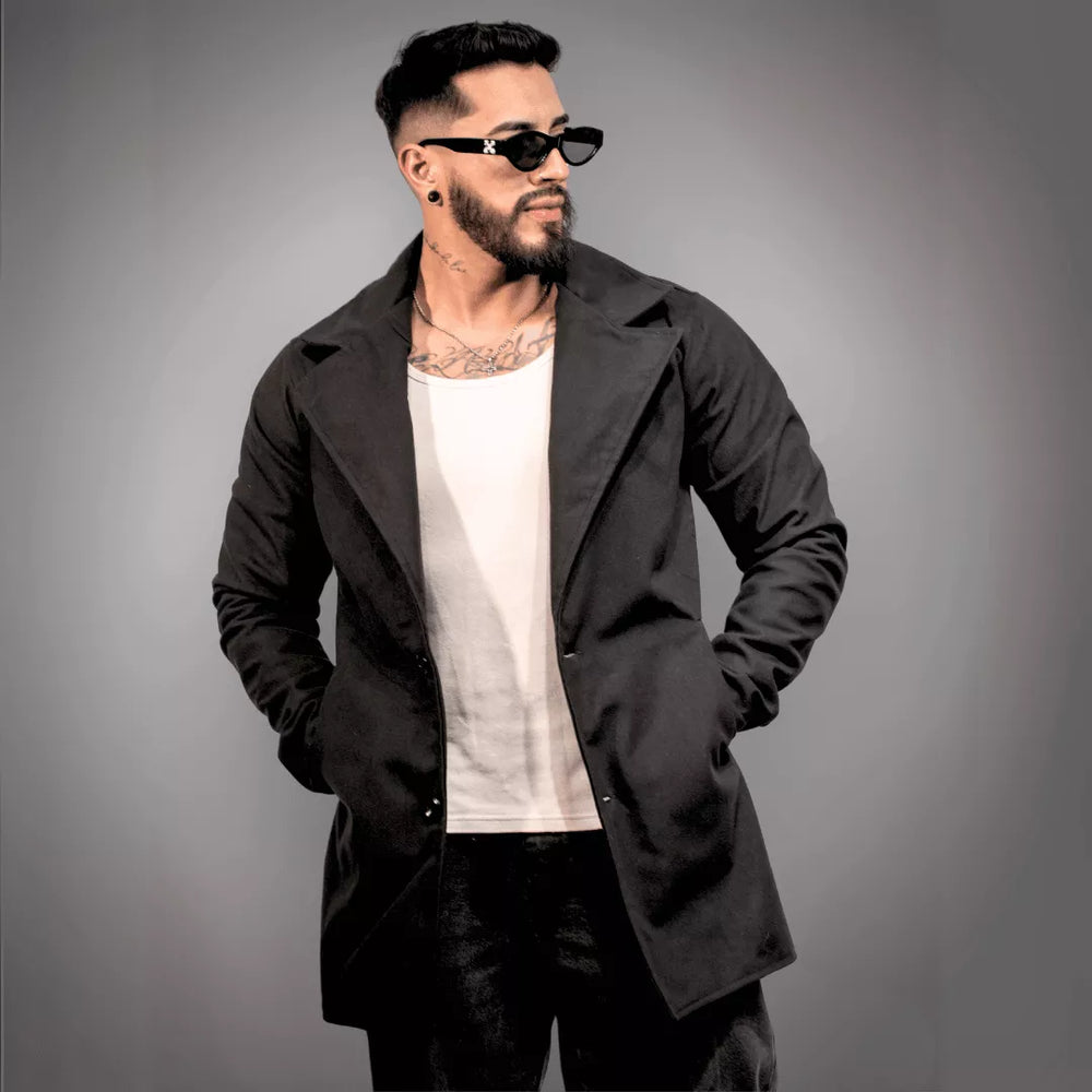 Gabardina negra clásica para hombre, versátil para cualquier ocasión. El modelo sonríe y posa con las manos en los bolsillos, con gafas de sol y camisilla blanca. Ideal para un look elegante o casual en Colombia