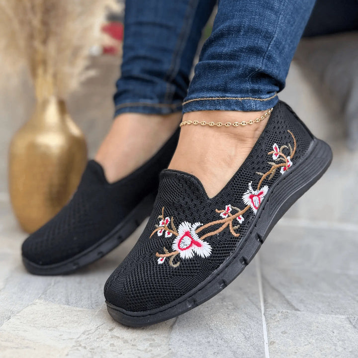Zapatillas Florenza negras con bordado floral en blanco y rosa, tejido knit transpirable, suela ligera antideslizante y diseño slip-on ergonómico para confort y estilo casual diario