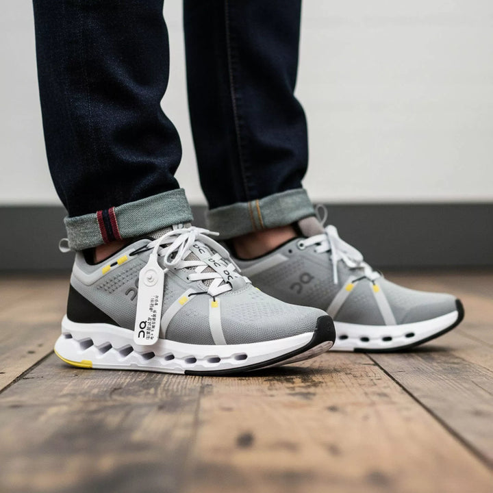 Primer plano de tenis On color gris con suela blanca y negra amortiguada, cordones blancos y marquilla de seguridad. Combinados con un jean oscuro con bota doblada sobre un piso de madera rústico