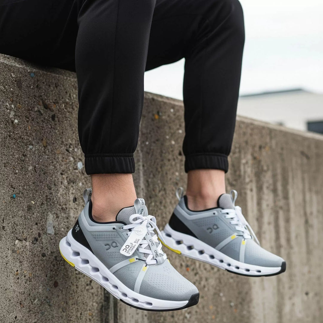 Persona sentada usando tenis On grises con suela blanca amortiguada y detalles amarillos. Combinados con un pantalón jogger negro con resorte en el tobillo, sobre un muro de concreto texturizado