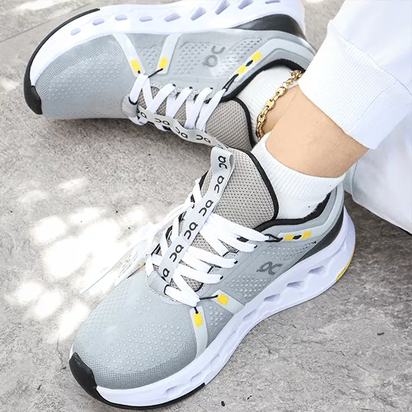 Outfit casual de dama con tenis marca On grises, pantalón blanco y un detalle de tobillera dorada. Una foto con iluminación natural que resalta el calzado deportivo premium en tendencia para mujeres.
