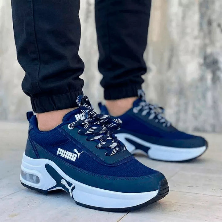 Tenis deportivos unisex de estilo robusto en color azul marino con plataforma blanca y cámara de aire visible. El calzado se apresenta en un modelo masculino con jogger negro, destacando un diseño moderno e imponente para el mercado colombiano