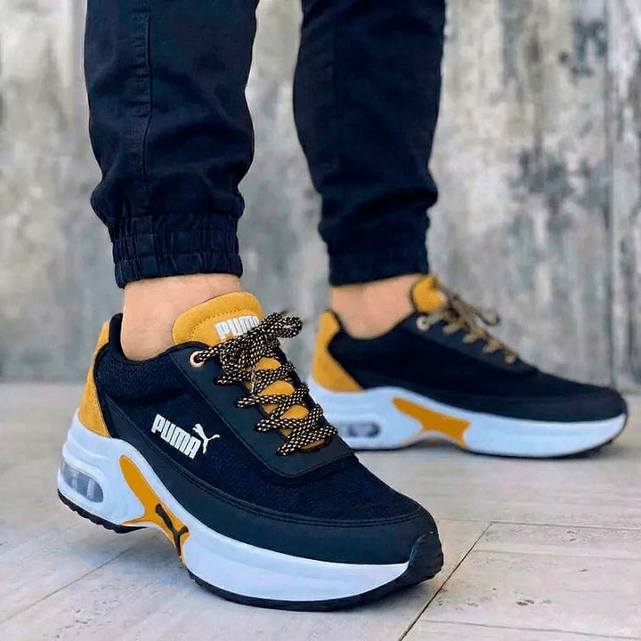 Tenis urbanos unisex negros con detalles vibrantes en color mostaza y entresuela blanca con cámara de aire. El modelo masculino viste jogger premium para enfatizar la estética audaz y el confort de este calzado exclusivo.