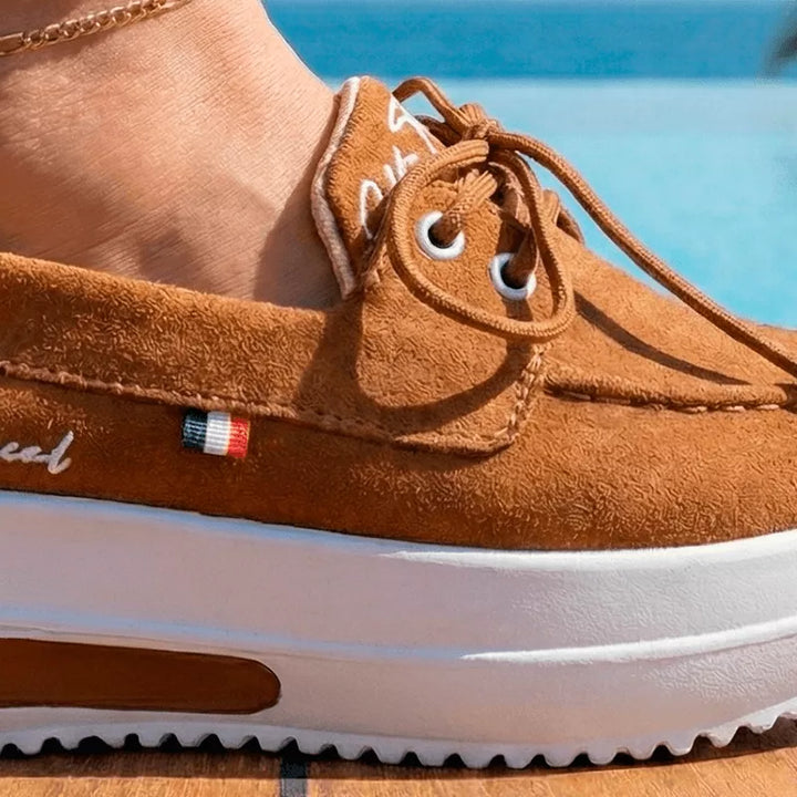 Zapatillas Francia color Miel en ambiente tropical de playa, pie sobre deck de madera con fondo de mar turquesa y palmeras. Calzado de verano cómodo.